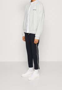 Veste bomber zippée gris clair sur un t-shirt blanc, associée à un pantalon bleu marine avec des bandes blanches sur les côtés et des baskets blanches.