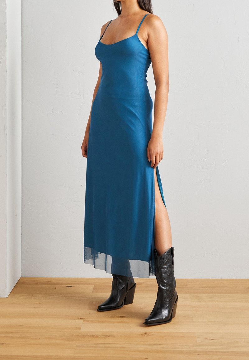 Femme portant une robe maxi bleue sans manches avec une fente latérale et des bottes noires à talons mi-mollet, debout sur un sol en bois.