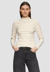 Rippstrick-Oberteil mit langen Ärmeln in Creme mit beigen horizontalen Streifen und hochgeschlossenem Kragen. Kombiniert mit dunkel schwarzen Jeans. Die Textur ist glatt.