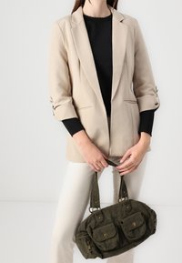 Béžové na míru šité sako přes černý top, doplněné krémovými kalhotami. V ruce drží zelenou manšestrovou kabelku se dvěma předními kapsami a bronzovým kováním.