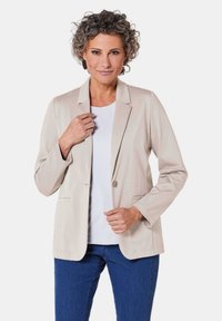 Beige blazer van zachte stof; heeft een enkele knoop, gesneden revers en voorzakken. Gecombineerd met een witte top en blauwe jeans.