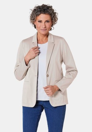 Beige Sakko aus weichem Stoff; verfügt über einen einzigen Knopf, Reverskragen und Vordertaschen. Kombiniert mit einem weißen Oberteil und blauen Jeans.