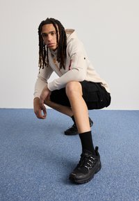 Felpa beige con tasca laterale con zip e dettaglio rosso, pantaloni cargo neri, sneakers neri e calzini neri al ginocchio su uno sfondo di moquette blu.