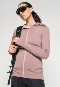 Moletom rosa com fecho de correr, capuz com cordão, punhos ribados e uma textura leve; emparelhado com óculos de sol pretos e uma alça de mochilas preta.