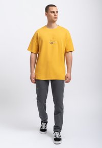 Geel katoenen T-shirt met een grafisch ontwerp op de voorkant, gecombineerd met grijze elastische broek met turquoise stiksels en zwarte sneakers.