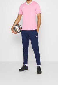 Maglietta da calcio rosa a maniche corte con scollo a V bianco, pantaloni tapered blu scuro, scarpe sportive nere e un pallone da calcio multicolore.