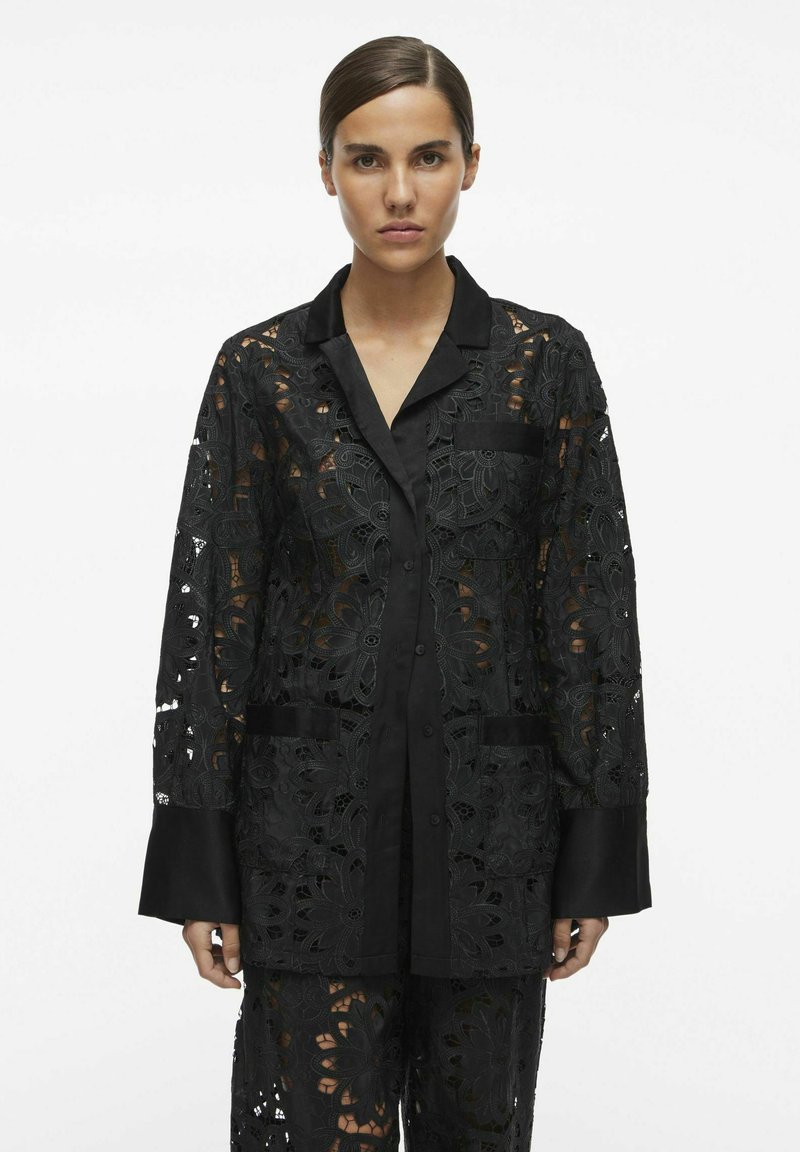 Veste en dentelle noire avec des motifs floraux découpés, des manches longues, un col et des poignets en satin noir, avec une fermeture à boutons et des poches avant.