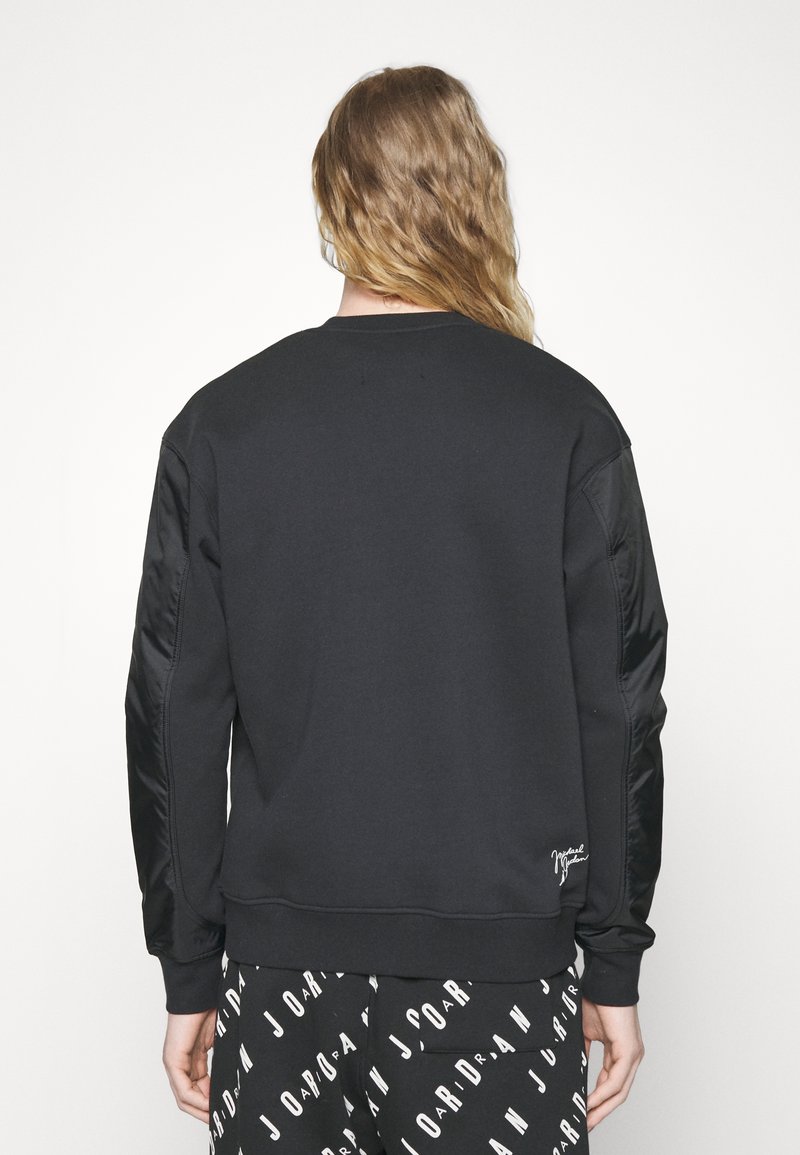 Sweat-shirt noir avec manches en satin, col rond et poignets côtelés. Présente un petit logo blanc à l'ourlet. Porté avec un pantalon noir imprimé.