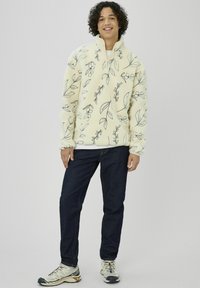 Pull polaire crème avec motifs de feuilles noirs, col montant, demi-fermeture éclair à l'avant. Jean en denim foncé et baskets grises avec des accents.