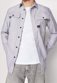 Vaaleanharmaa nappipaita, jossa on kaksi rintataskua, mustat napit ja musta merkki. Päällä valkoinen paita ja harmaat farkut.