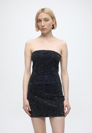 Γυναίκα με κοντά ξανθά μαλλιά που φοράει strapless μαύρο μίνι φόρεμα με λευκές πουά, όρθια μπροστά σε απλό λευκό φόντο.