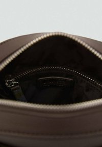 Interno di una borsa in pelle marrone che mostra una texture liscia, un compartimento con zip e un'etichetta attaccata sul lato. Dettaglio della zip argentata visibile.