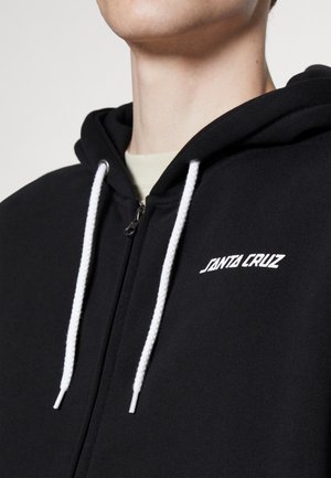 Nahaufnahme einer Person, die einen schwarzen Kapuzenpullover mit weißen Kordeln und "Santa Cruz" auf der Brust trägt.