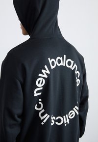 Hoodie noir avec un logo blanc arrondi au dos. Texture lisse et poche kangourou. Présente une coupe décontractée et une capuche.