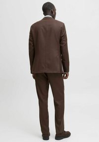 Brauner Anzug mit maßgeschneidertem Design, bestehend aus einem Blazer mit einem Schlitz und passenden Hosen. Glatte Textur, einfache Stecknähte.