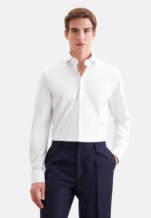 Jeune homme portant une chemise blanche boutonnée et un pantalon bleu marine foncé, debout devant un fond clair uni.