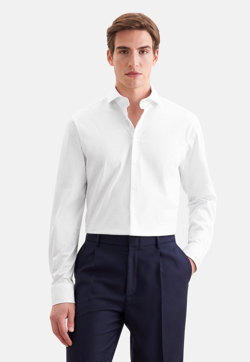 Jeune homme portant une chemise blanche boutonnée et un pantalon bleu marine foncé, debout devant un fond clair uni.
