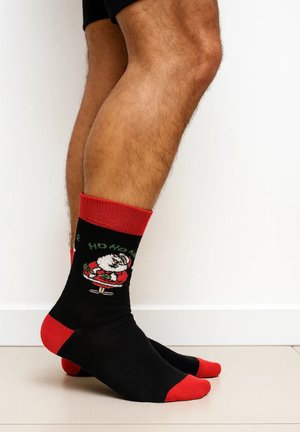 Schwarze Socken mit roten Akzenten, die eine Weihnachtsmann-Grafik und den Schriftzug "Ho Ho Ho" zeigen. Hergestellt aus weichem Material. Knöchellanges Design, gerippter Bund.