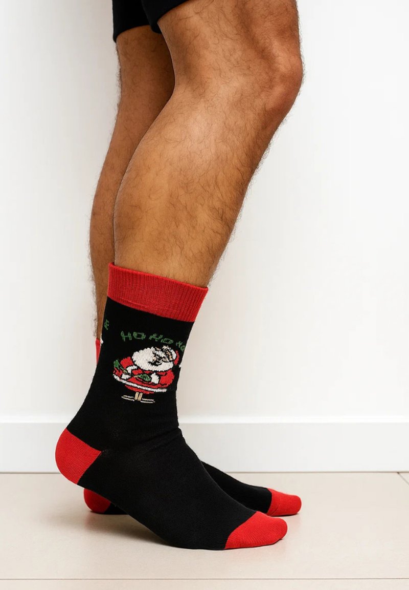 Schwarze Socken mit roten Akzenten, die eine Weihnachtsmann-Grafik und den Schriftzug "Ho Ho Ho" zeigen. Hergestellt aus weichem Material. Knöchellanges Design, gerippter Bund.