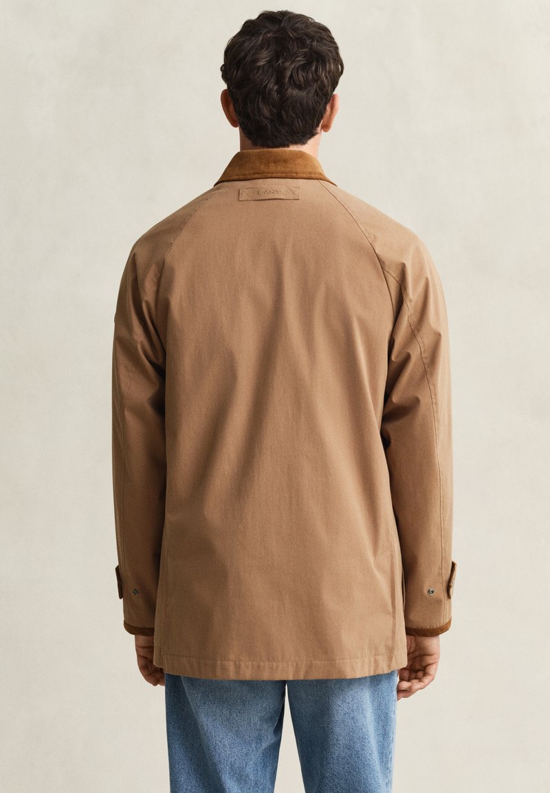 Homme portant une veste marron avec un col en velours côtelé et un jean bleu, vu de dos sur un fond clair uni.