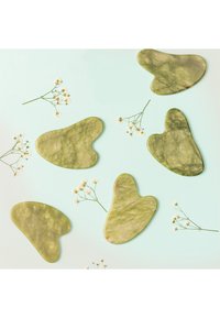 Grüne Jade-Gesichtsmassagetools mit organischen Formen, glatter Textur und marmorierten Mustern, angeordnet zwischen kleinen weißen Blumen auf einem hellen Hintergrund.