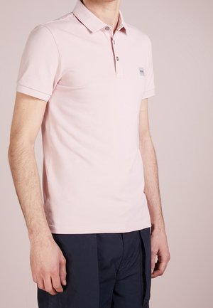 Mann trägt ein hellrosa kurzärmeliges Poloshirt mit Knöpfen und marineblaue Hose, steht vor einem schlichten beigen Hintergrund.