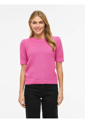 Kobieta o długich blond włosach, ubrana w różowy, teksturowany sweter z krótkim rękawem i czarne spodnie, uśmiechająca się i stojąca na białym tle.