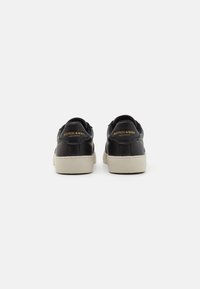 Adidași din piele neagră cu o textură netedă, cu vârf rotund, talpă din cauciuc alb și logo auriu pe călcâi. Design simplu la spate.