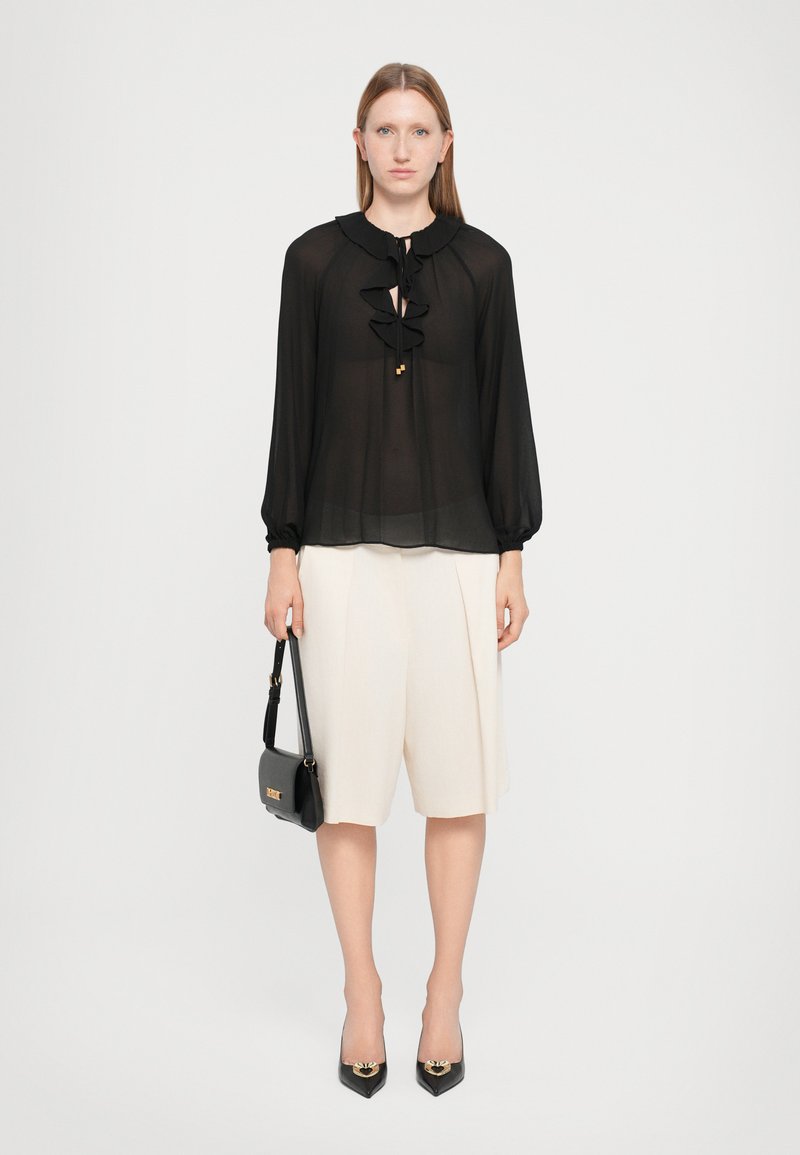 Black sheer blouse with ruffled front, keyhole neck, and long sleeves. Paired with cream pleated shorts and black pointed-toe heels.