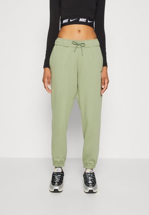 Femme portant un pantalon de jogging vert clair, un crop top noir avec le logo Nike, et des baskets Nike noires et blanches, debout devant un fond uni.