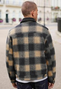 Flanneljakke med et sort og beige ternet mønster, der har en krave og trykknapper. Blød tekstur og afslappet pasform, mellemlang design.