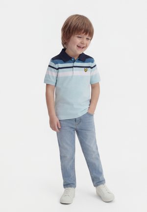 Băiat tânăr zâmbind, purtând un tricou polo cu dungi albastre, blugi deschiși, adidași albi, stând cu o mână în buzunar pe un fundal alb.