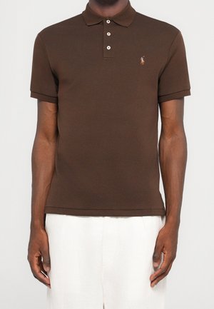 Homme portant un polo marron ajusté avec un petit logo brodé sur la poitrine, associé à un pantalon blanc large.