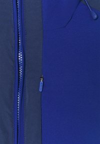Veste zippée bleue présentant un design en tissu mixte avec des textures contrastées, une fermeture éclair lisse avec un tirette bleue, et une coupe près du corps.