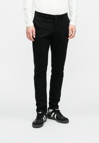 Černé úzké kalhoty s hladkou texturou, se zapínáním na knoflík vpředu a bočními kapsami, v kombinaci s černými teniskami.