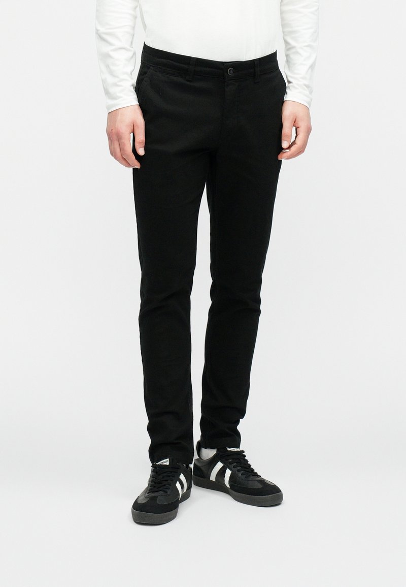 Černé úzké kalhoty s hladkou texturou, se zapínáním na knoflík vpředu a bočními kapsami, v kombinaci s černými teniskami.