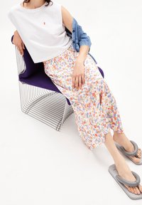 Kvinna som sitter på en trådstolsstol iförd en ärmlös vit topp, blommiga vida byxor, jeansjacka och grå vadderade sandaler.