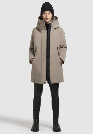 Parka impermeabile beige con colletto alto, tasche con zip e orlo regolabile, abbinata a leggings neri aderenti e stivali al ginocchio.