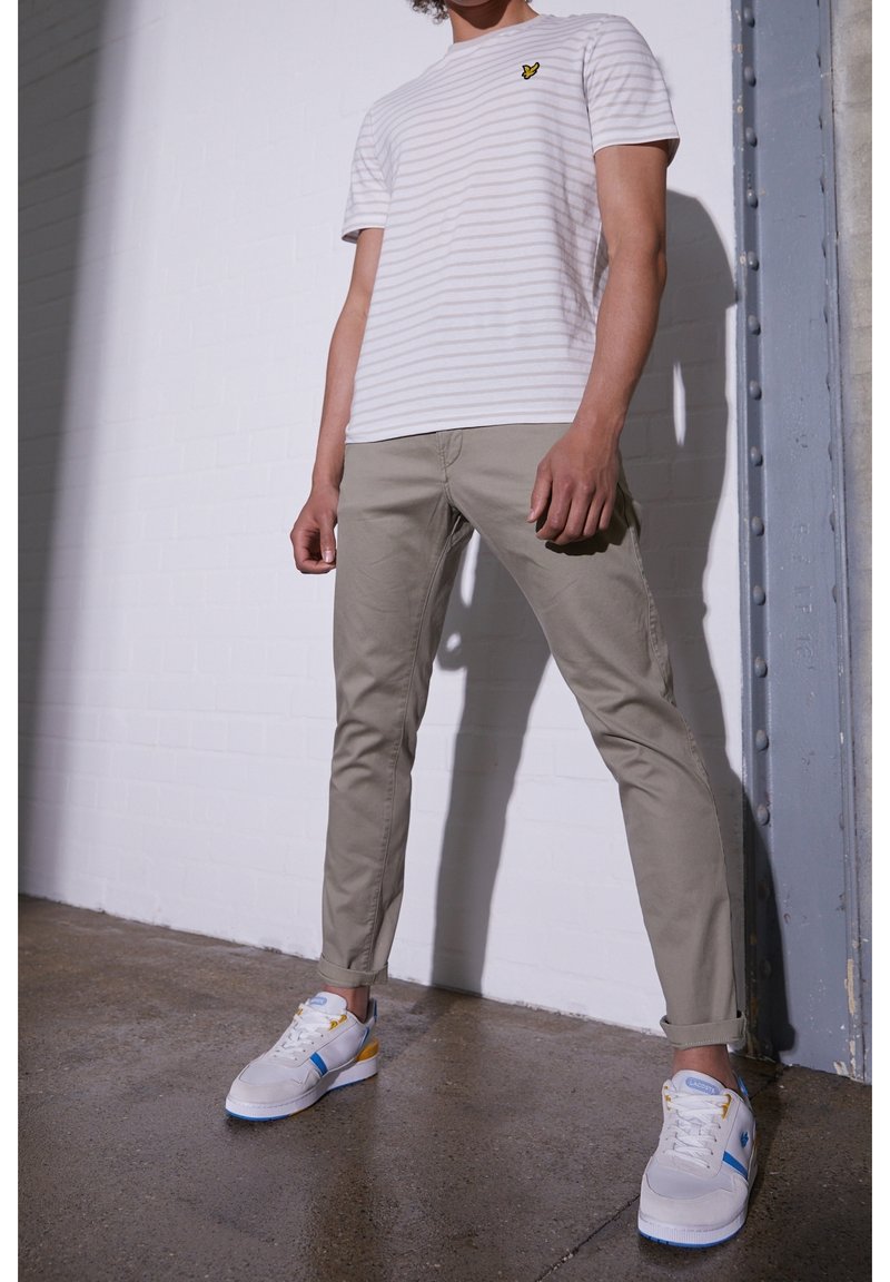 Camiseta de manga corta blanca a rayas, pantalones beige ajustados y zapatillas blancas con detalles en azul y amarillo. Entorno industrial con un suelo texturizado.