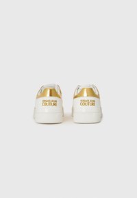 Sneakers brancos com um salto metálico dourado, apresentando a marca "VERSACE JEANS COUTURE" em relevo na parte de trás e uma textura suave.