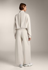 Kermanvärinen, teksturoidun ilmeen vaatekokonaisuus, joka sisältää korkeakauluksisen paidan, jossa on takatasku, ja leveälahkeiset housut. Korostettu vaaleanharmailla kengillä.