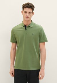 Zelená bavlnená polo košeľa s golierom, trojicou gombíkov a malým logom na hrudi, v kombinácii s tmavými nohavicami.
