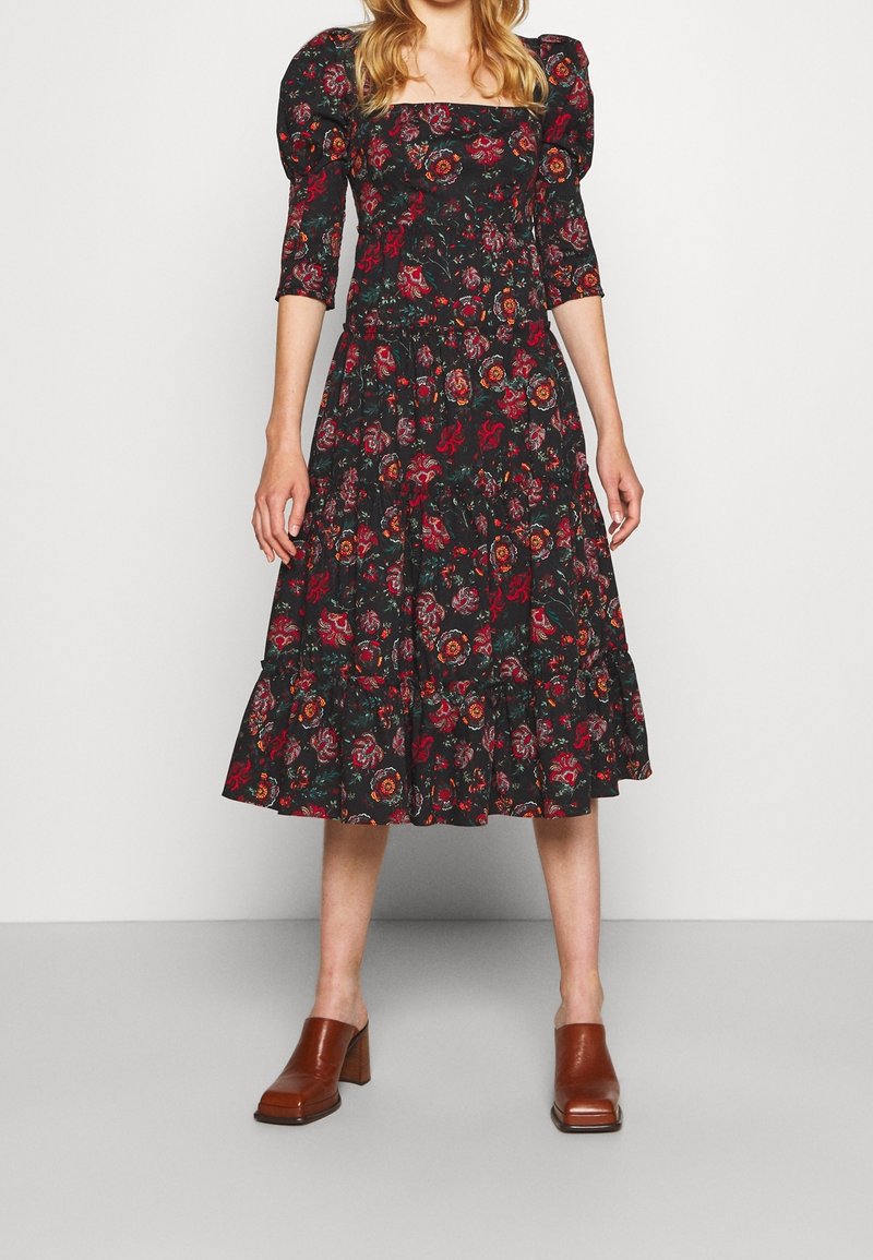 Femme portant une robe midi noire à fleurs avec des manches bouffantes et des mules marron à talons blocs, debout devant un fond clair uni.
