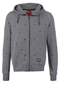 Szara bluza z kapturem zapinana na suwak, z czarnymi wzorami gwiazd, dwoma przednimi kieszeniami i kapturem ze sznurkiem. Posiada oznakowaną etykietę w pobliżu kieszeni.