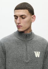 Gray knit sweater with a high collar, half-zip, and textured fabric, featuring a cream "W" logo on the left chest.
