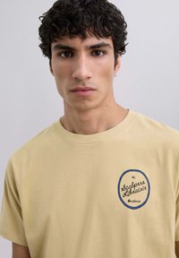 Camiseta de color beige claro hecha de algodón, con un logo redondo bordeado en azul en el lado izquierdo del pecho que dice "la Librairie Scalpers Bordeaux."