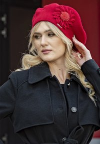 Czerwona beretka z teksturowanej tkaniny, ozdobiona kwiatowym akcentem z koralikami. Modelka nosi czarny płaszcz z szerokim, wykładanym kołnierzem.