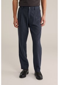 Bărbat purtând pantaloni plisati bleumarin închis și loaferi negri, stând pe un fundal bej simplu.