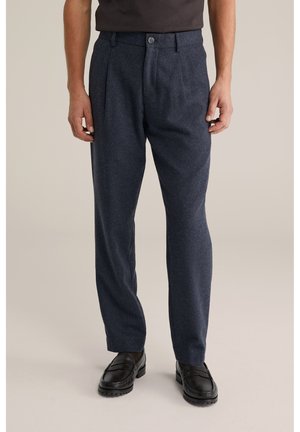 Muž oblečený v tmavomodrých plisovaných nohaviciach s čiernymi mokasínami, stojaci pred jednoduchým béžovým pozadím.