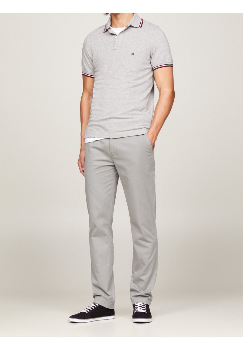 Homme portant un polo gris clair avec col et manches rayés, pantalon gris clair, baskets noires avec lacets blancs, debout avec une main dans la poche.